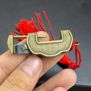 May include: A brass-colored metal padlock with an open U-shaped design and engraved Chinese characters on the front. The lock features a sliding mechanism and is attached to a bright red decorative tassel made of thin, dangling fabric cords.