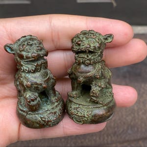 Peut inclure: Deux petites figurines en métal de couleur bronze patiné représentant des lions gardiens, avec des détails sculptés complexes, une expression aux yeux écarquillés et une base arrondie ornée de motifs en relief, présentant une oxydation verte.