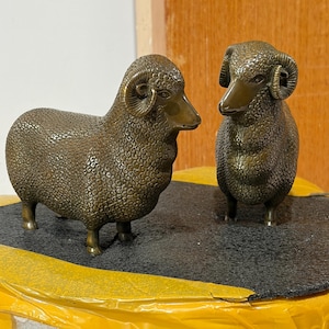 Puede incluir: Dos figuras de carnero de color bronce con patrones de lana detallados y texturizados y cuernos curvados. Los carneros están representados en posición de pie, uno mirando hacia la derecha y el otro hacia adelante, con un acabado metálico.