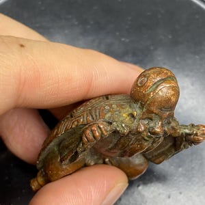 May include: A small, detailed bronze turtle figurine. The turtle has a textured shell and head, with visible legs and a tail. The bronze has a patina, showing age and wear. The figurine is held in a person's hand.