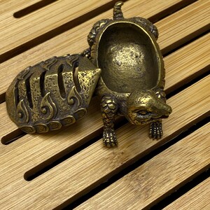 May include: A detailed brass turtle-shaped container with an open top and a hinged shell. The container has intricate details, including textured scales and a small tail. The turtle is positioned on a wooden surface with parallel grooves.