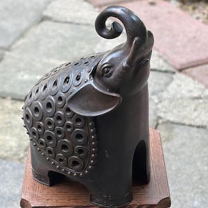 May include: A dark brown metal elephant figurine with a trunk curled upward. The body features a decorative pattern of raised circular rings across the back and sides, with a beaded border detail along the edge of the saddle-like section.