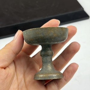 May include: A small, weathered brass chalice featuring a wide, shallow bowl atop a slender, rounded stem and a circular base. The metal surface displays a dark, aged patina with visible golden-toned highlights throughout the entire structure.