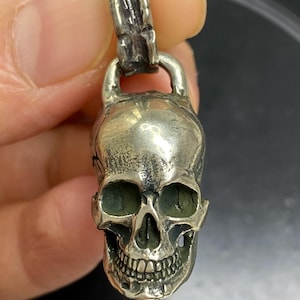 May include: Silver skull pendant with detailed facial features, including eye sockets and teeth. The pendant has a bail for attaching to a chain. The skull is a classic symbol often associated with gothic or biker aesthetics.