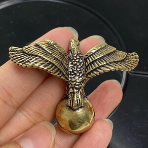 May include: A detailed brass eagle figurine with outstretched wings, perched atop a spherical base. The eagle's feathers are intricately carved, showcasing a vintage aesthetic. The sculpture is held in a hand, highlighting its size and detail.
