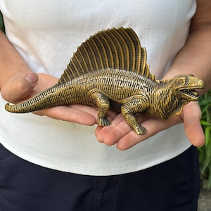 Op de afbeelding: Een metalen, bronskleurig beeldje van een Dimetrodon, een prehistorische synapside, met een opvallend zeil op de rug, textuurdetails op de huid en een geopende bek met zichtbare tanden, horizontaal vastgehouden met de staart naar links.