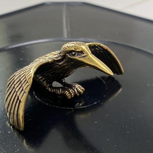 May include: A detailed bronze-colored bird figurine with outstretched wings and a long beak. The bird is perched on a dark surface, showcasing intricate feather and claw details. The figurine has a vintage or antique aesthetic.