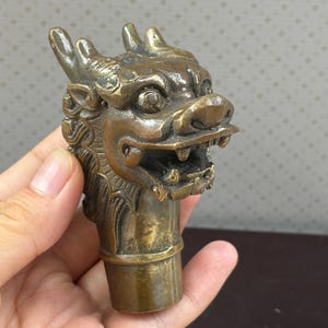 Può includere: Una scultura in metallo color bronzo a forma di testa di drago con squame intricate, corna prominenti e una bocca aperta con denti visibili. La scultura è fissata su una base cilindrica e presenta una finitura metallica usurata con patina.