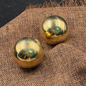 Pode incluir: Duas esferas metálicas de cor dourada polida com uma superfície lisa e refletora. Cada esfera tem um acabamento de alto brilho que cria um efeito de espelho, mostrando um reflexo claro do ambiente no seu exterior curvo.
