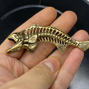 May include: A detailed, antique-style brass fish skeleton figurine. The open-mouthed fish has visible ribs, fins, and tail, with a textured, aged appearance. The figurine is a decorative item, possibly a pendant or charm.