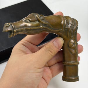 May include: A bronze-colored metal cane handle featuring a sculpted horse head with a bridle, flowing mane, and a circular emblem on the side. The handle has a cylindrical base designed to attach to a walking stick shaft with a smooth finish.