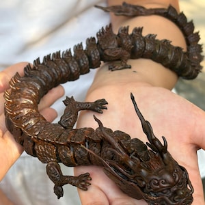 May include: A dark brown articulated dragon figurine with a segmented, flexible body, detailed scales, and a stylized head featuring long, sweeping horns. The creature has four clawed limbs and a long, winding tail that curves across the palm.