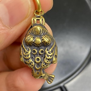 Può includere: Un piccolo ciondolo in ottone in stile antico con un design dettagliato. Il ciondolo presenta una figura stilizzata di animale con motivi intricati e un anello per appenderlo. Il colore generale è un ottone caldo e invecchiato.