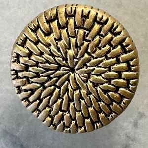 May include: A circular cabinet knob in an antique brass finish, featuring a woven texture. The knob's design is a detailed pattern that spreads out from the centre. The finish has a slightly aged appearance.