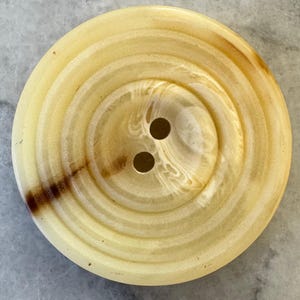 Könnte beinhalten: Ein großer, runder Knopf mit einem hellgelben und braunen Wirbelmuster. Der Knopf hat zwei Löcher in der Mitte und eine glänzende Oberfläche. Der Knopf ist ein Näh- oder Bastelzubehör.