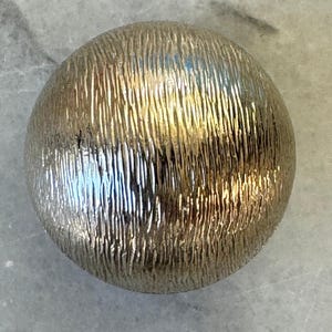 Könnte beinhalten: Ein kugelförmiges Objekt mit metallisch goldener Oberfläche und einer tief texturierten, vertikal gestreiften Struktur. Die Kugel reflektiert das Licht auf ihrer gewölbten Außenseite und betont die feinen, linearen Rillen auf der Oberfläche.