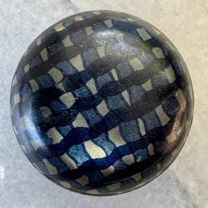 Puede incluir: Un objeto decorativo redondo con un patrón de cuadros azul oscuro y dorado. El objeto tiene un acabado brillante y está sobre un fondo gris claro. El diseño presenta una disposición en forma de cuadrícula de formas azules y doradas.