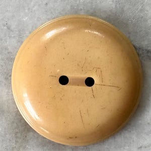以下が含まれることがあります： 2つの黒い穴がある、大きくて丸い薄いタン色のボタン。ボタンはわずかに光沢があり、傷が見えます。このヴィンテージボタンは、縫製プロジェクトや工芸品に適しています。