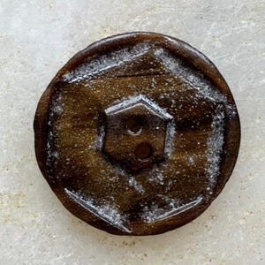 May include: A circular brown button featuring a central hexagonal recessed area with two holes for attachment. The surface of the button displays a dark, polished finish with subtle, light-colored speckling and a distinct geometric design.