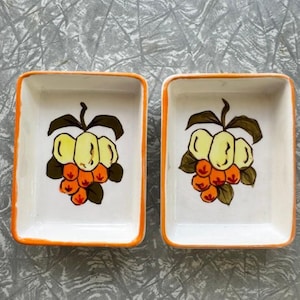 Könnte beinhalten: Zwei rechteckige Keramikschalen mit orangefarbenem Rand. Jede Schale zeigt ein handgemaltes Design aus einer Ansammlung gelber und orangefarbener Früchte mit grünen Blättern. Die Schalen stehen vor einem grauen Hintergrund.