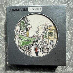 May include: A set of ceramic tile coasters in a grey box. The coasters display a detailed illustration of a street scene with people, buildings, and a green awning. The box is labelled "CERAMIC TILE" and "COASTERS".