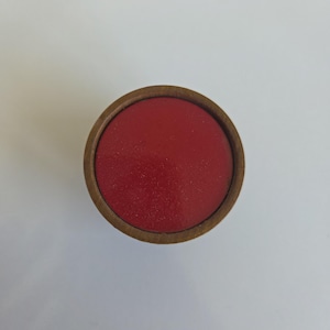 May include: A circular object featuring a smooth, solid red top surface enclosed within a natural brown wooden rim. The object is viewed from directly above, highlighting the contrast between the vibrant red center and the circular wood frame.