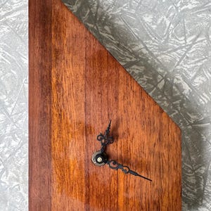 May include: A wooden clock with a unique, angled design. The clock face is made of rich, dark brown wood with vertical grain. Black clock hands and a central mechanism complete the minimalist aesthetic. The clock measures approximately 30 cm tall.