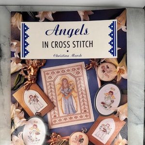 May include: A book titled "Angels in Cross Stitch" by Christina Marsh. The cover features cross-stitch angel designs in various frames and a central angel image. The color palette includes gold, blue, and cream.