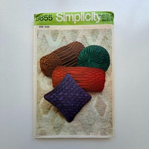 May include: Simplicity pattern 9855 showing decorative pillows in brown, green, red, and purple. The pillows have textured and pleated designs. The pattern envelope is green and white.