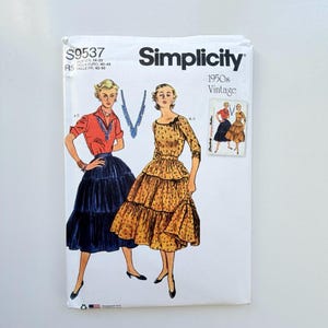 May include: A Simplicity S9537 sewing pattern envelope for 1950s vintage style clothing. The white paper envelope features illustrations of two outfits: a red blouse with a dark blue tiered skirt, and a yellow floral print dress with a tiered skirt.