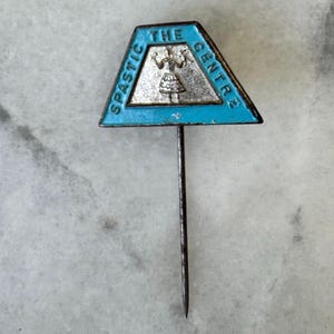 Könnte beinhalten: Vintage Anstecknadel mit einem dreieckigen, blau emaillierten Oberteil und einer silbernen Figur. Der Text "SPASTIC CENTRE" und "THE" und "CENTRE" sind sichtbar. Die Nadel hat eine lange, dünne Metallnadel.