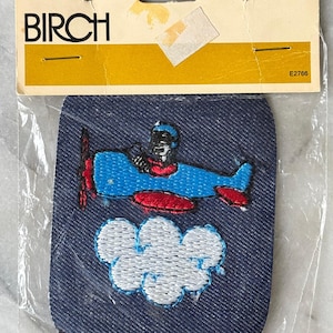 May include: A denim patch featuring a blue airplane with a red propeller and accents, a pilot, and a white cloud. The patch is packaged with a cardboard header that reads "BIRCH" and the number "E2766".