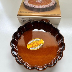 May include: Brown stoneware quiche pan with a scalloped edge, and a matching box. The box reads "Stoneware Oven Cookware Quiche Pan". The pan has a glossy finish and a sticker that reads "Oven to Table Stoneware, Also Suitable for Microwave Ovens, Dishwasher Safe."