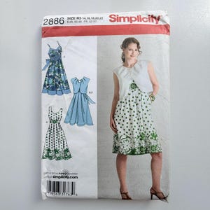 Könnte beinhalten: Simplicity Schnittmuster 2886 für die Größen 14-22. Die Hülle zeigt Illustrationen von drei Kleider-Varianten und einer Bolero-Jacke sowie ein Foto eines Modells, das einen weißen Bolero über einem grün-weiß gepunkteten Blumenkleid trägt.