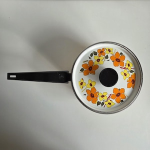 May include: A round metal saucepan with a white lid featuring a floral pattern of orange and yellow flowers with green leaves. The pan has a long, straight black handle and a central black knob on the lid, viewed from directly above.