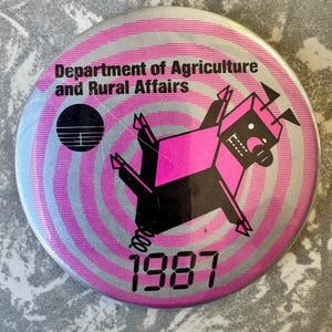 May include: A circular badge with a pink and silver spiral pattern. The text "Department of Agriculture and Rural Affairs" and the year "1987" are displayed. A pink and black cartoon character is also featured.
