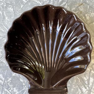 May include: A dark brown, shell-shaped ceramic dish. The dish has a scalloped edge and a textured, ridged surface, with a small rectangular base. The glossy finish reflects light, highlighting the details of the design. This decorative piece is suitable for serving or display.