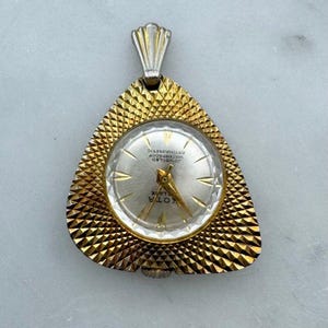May include: A vintage gold-toned pendant watch with a triangular frame and a silver-colored watch face. The watch face features gold hands and the word "KOTA". The watch is marked "JEWELED WATERPROOF ANTIMAGNETIC".