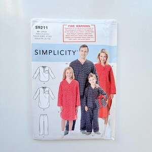 Op de afbeelding: Een Simplicity naaipatroon envelop S9211 voor kindernachtkleding. De verpakking toont illustraties van een top met lange mouwen en een broek, naast foto's van drie personen die rood bedrukte en marineblauw geruite pyjama's dragen.