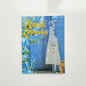 以下が含まれることがあります： Family Circle Romantic Embroidery: Practical Needlework Projects to Make and Treasureというタイトルのソフトカバー本。表紙には、緑の葉が茂る鮮やかな青い木製のドアに掛けられた、黒い刺繍が施された白いエプロンが写っています。
