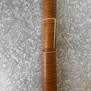 May include: A cylindrical roll of adhesive contact paper featuring a realistic medium brown wood grain pattern. The roll is partially unspooled, revealing a white backing layer along the edge of the wood-textured surface of the material.