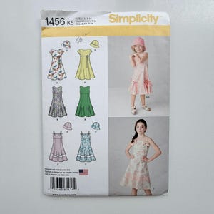 May include: Simplicity sewing pattern 1456 K5 for sizes 7-14. The envelope features illustrations of various dress styles with matching hats, including floral, solid, and patterned designs. Two photographs show models wearing the finished dresses.