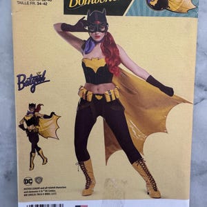May include: Simplicity sewing pattern featuring a DC Comics Bombshells Batgirl costume. The pattern includes sizes U.S. 6-14 and Euro 32-40. The cover art showcases a Batgirl illustration with a yellow cape and black outfit.