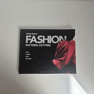 以下が含まれることがあります： Zarida Zaman著、Bloomsbury出版の「Fashion Pattern Cutting」というタイトルの長方形の本。黒い表紙には白い文字と、ギャザーの入った赤い衣服の写真が印刷されており、デザインにおけるライン、シェイプ、ボリュームが強調されています。