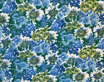 May include: A close-up of a fabric with a floral pattern. The design features a dense arrangement of blue, light blue, and green flowers against a white background. The fabric has a textured appearance, suggesting a woven material.