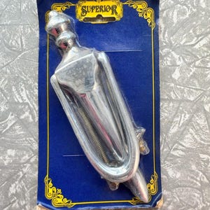 May include: A chrome-colored door knocker in its original packaging. The knocker is shaped like a stylized figure with a curved handle and a spring mechanism. The packaging is blue with gold decorative trim and the word "SUPERIOR".