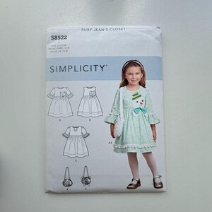 May include: Simplicity sewing pattern S8522 featuring a child's dress with a snowman applique, a matching purse, and other dress designs. The pattern includes sizes US 3-8.