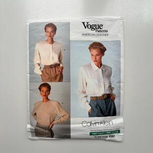 May include: A Vogue Patterns American Designer sewing pattern envelope for Calvin Klein, number 2397. The white paper envelope features three photographs of long-sleeved shirts with collars or crew necks, paired with skirts or trousers, labelled A, B, and C.