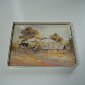 May include: A set of rectangular coasters featuring a watercolor painting of a rural house with a porch, trees, and a dirt road. The artwork is in a muted color palette of beige, brown, and green, presented in a white box.