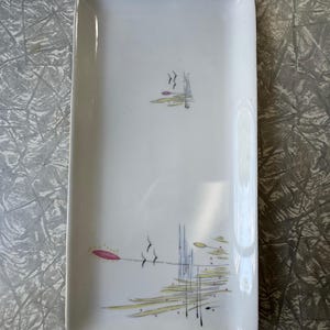 Könnte beinhalten: Ein rechteckiges weißes Keramik-Serviertablett mit abgerundeten Ecken. Das Tablett hat ein modernes Design aus der Mitte des Jahrhunderts mit abstrakten floralen und Wasserrohrmotiven in Rosa, Gelb und Schwarz. Das Tablett ist etwa 25 cm lang.